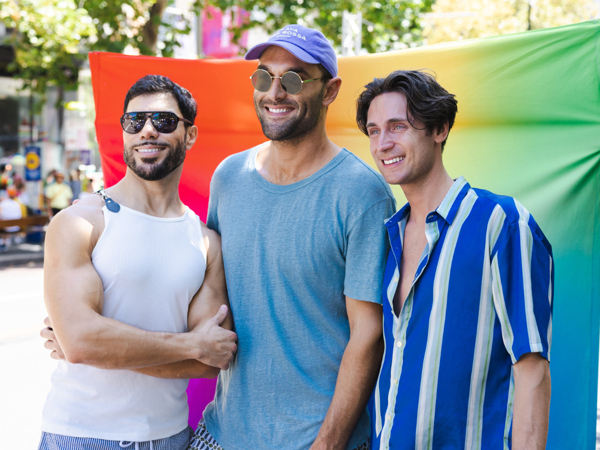 Group of men smiling in front of a rainbow backdrop at a Pride festival in the sun